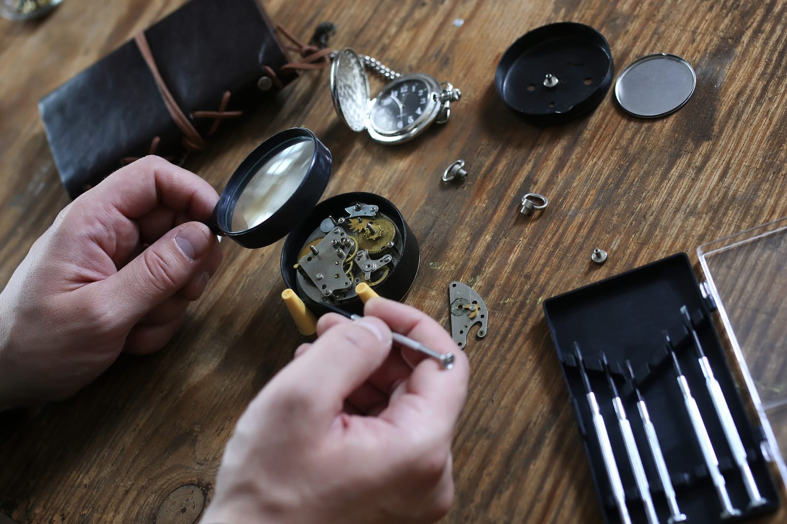 Orologiaio al banco che ispeziona con una lente un orologio da tasca smontato, con cacciaviti e vassoio di attrezzi accanto